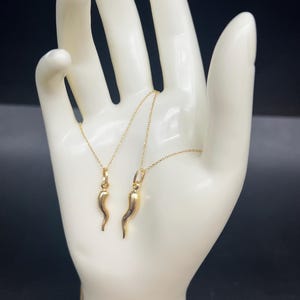 May include: Two gold necklaces with a horn charm pendant. The necklaces are displayed on a white hand-shaped stand against a black background.