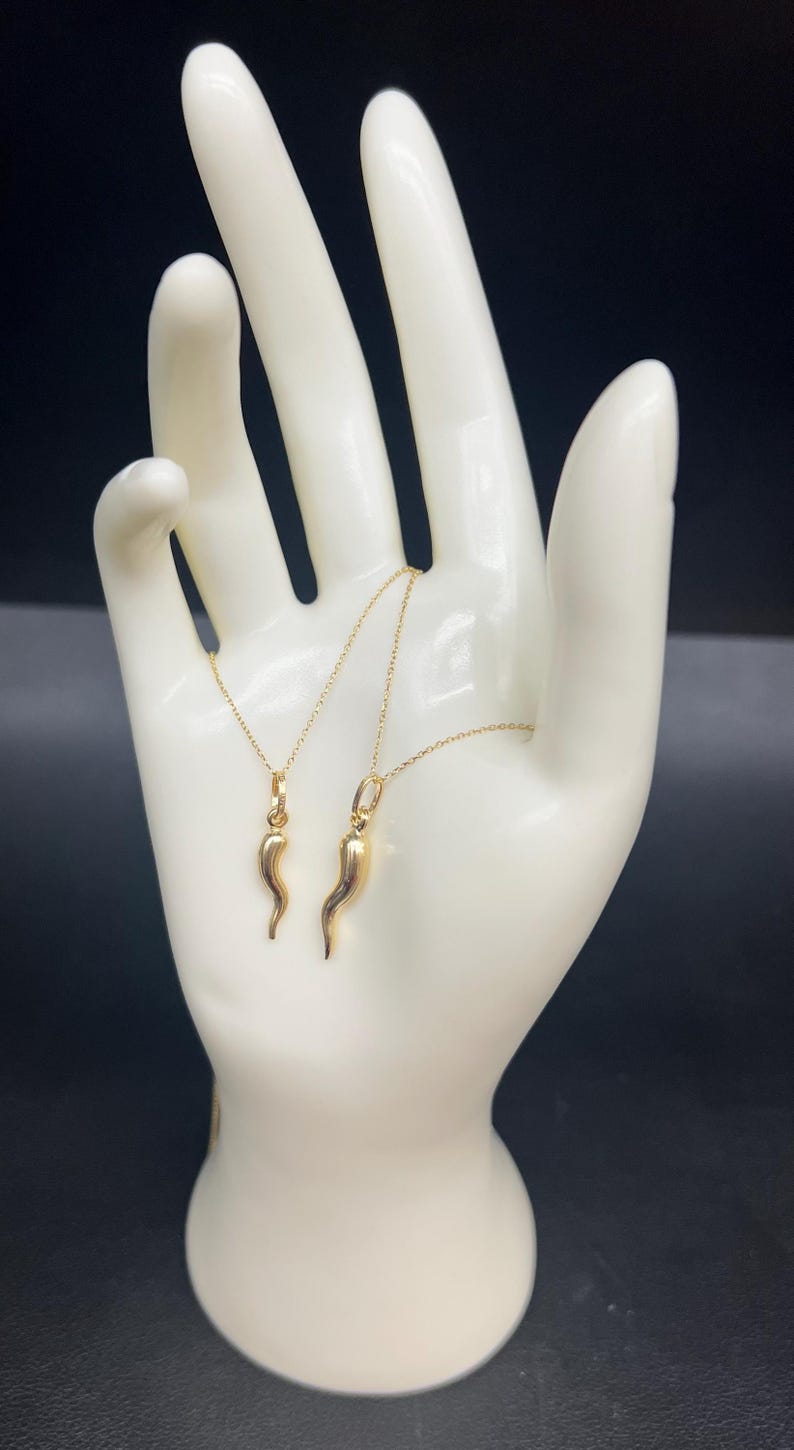 May include: Two gold necklaces with a horn charm pendant. The necklaces are displayed on a white hand-shaped stand against a black background.