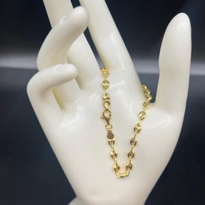 K&ouml;nnte beinhalten: Ein goldenes Kettenarmband mit einem zarten, runden Gliederdesign. Das Armband wird auf einer wei&szlig;en Mannequinhand pr&auml;sentiert.