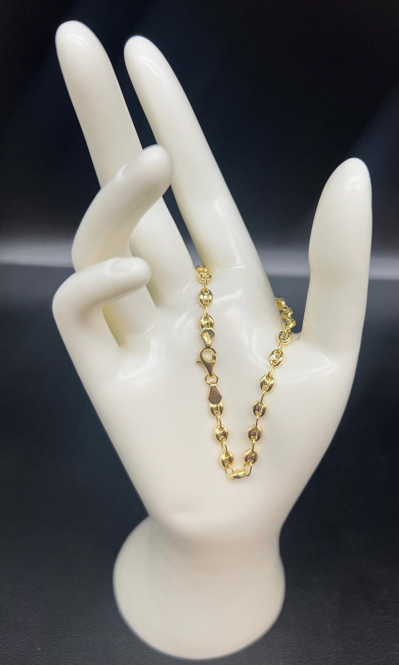 K&ouml;nnte beinhalten: Ein goldenes Kettenarmband mit einem zarten, runden Gliederdesign. Das Armband wird auf einer wei&szlig;en Mannequinhand pr&auml;sentiert.