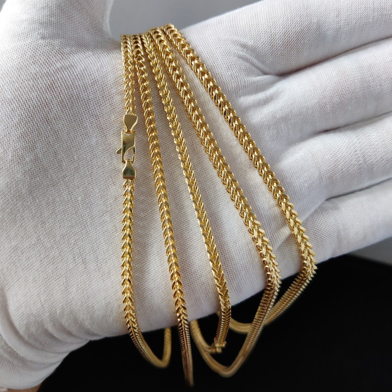 May include: Five gold chains of different lengths and widths, all with a herringbone pattern, are displayed on a white surface.