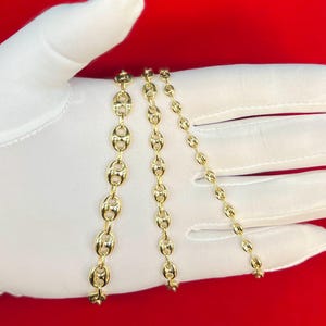 K&ouml;nnte beinhalten: Drei goldene Ketten mit einem Ankerkettenmuster. Die Ketten sind unterschiedlich lang und werden auf einer wei&szlig;en Oberfl&auml;che gezeigt.