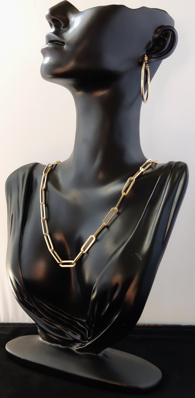 May include: A gold chain necklace with a link style and a pair of gold hoop earrings. The jewelry is displayed on a black mannequin bust.