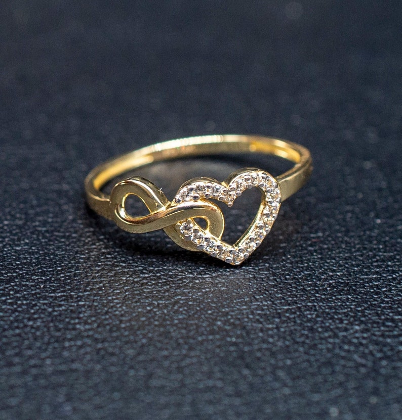 May include: A gold ring with a heart and infinity symbol design. The heart is outlined with small clear stones.
