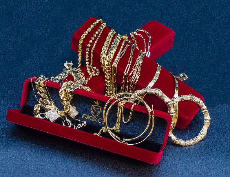 Puede incluir: Una colecci&oacute;n de cadenas, pulseras y pendientes de oro expuestos en cajas de terciopelo rojo. Las joyas est&aacute;n dispuestas de forma decorativa, mostrando los intrincados dise&ntilde;os y la artesan&iacute;a. Las cajas llevan la inscripci&oacute;n "Josh's Gold" y las iniciales "BG".