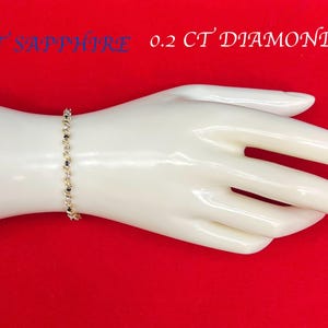 May include: A gold bracelet with a chain of alternating round and square links. The bracelet is on a white mannequin hand against a red background. The text "1 CT SAPPHIRE 0.2 CT DIAMOND" is printed on the background.