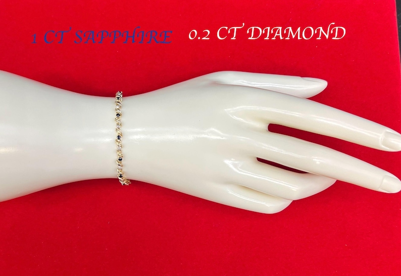 May include: A gold bracelet with a chain of alternating round and square links. The bracelet is on a white mannequin hand against a red background. The text "1 CT SAPPHIRE 0.2 CT DIAMOND" is printed on the background.