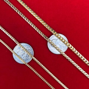 May include: Three gold chains of different widths on a red background. The chains are laid out in a diagonal pattern, with the widest chain at the top and the narrowest chain at the bottom. A silver coin is placed under each chain.