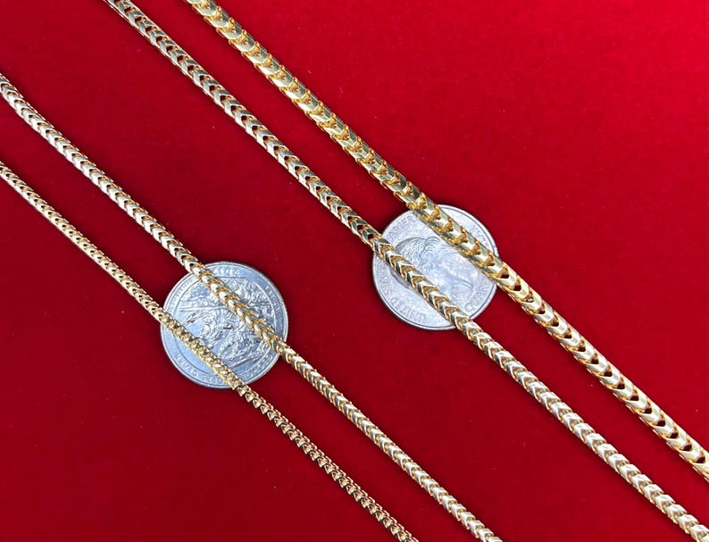 May include: Three gold chains of different widths on a red background. The chains are laid out in a diagonal pattern, with the widest chain at the top and the narrowest chain at the bottom. A silver coin is placed under each chain.