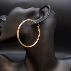May include: Gold hoop earrings on a black mannequin head. The earrings are large and circular.