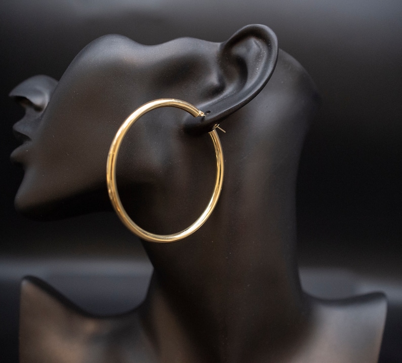May include: Gold hoop earrings on a black mannequin head. The earrings are large and circular.