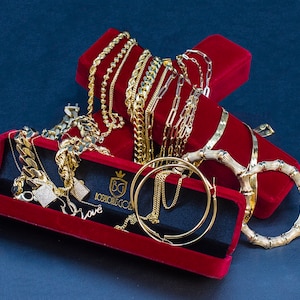 May include: An assortment of gold jewelry, including necklaces, chains, and hoop earrings, displayed in a red velvet jewelry box. The jewelry box has the text "Bosphorus Gold" on it. The jewelry is set against a dark blue background.