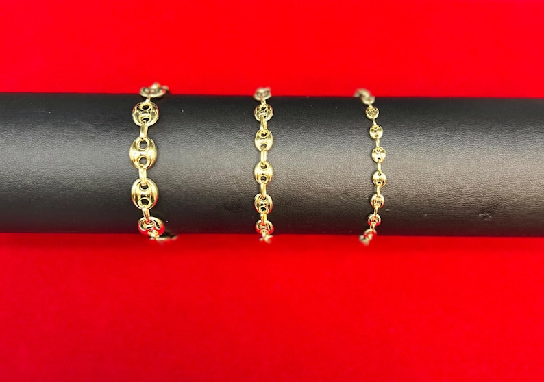 K&ouml;nnte beinhalten: Drei goldene Kettenarmb&auml;nder in verschiedenen L&auml;ngen, auf einer schwarzen Oberfl&auml;che pr&auml;sentiert.