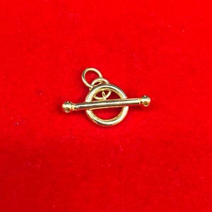 May include: A gold-colored toggle clasp charm, featuring a circular ring with a horizontal bar and two rounded ends. The charm is set against a vibrant red background.