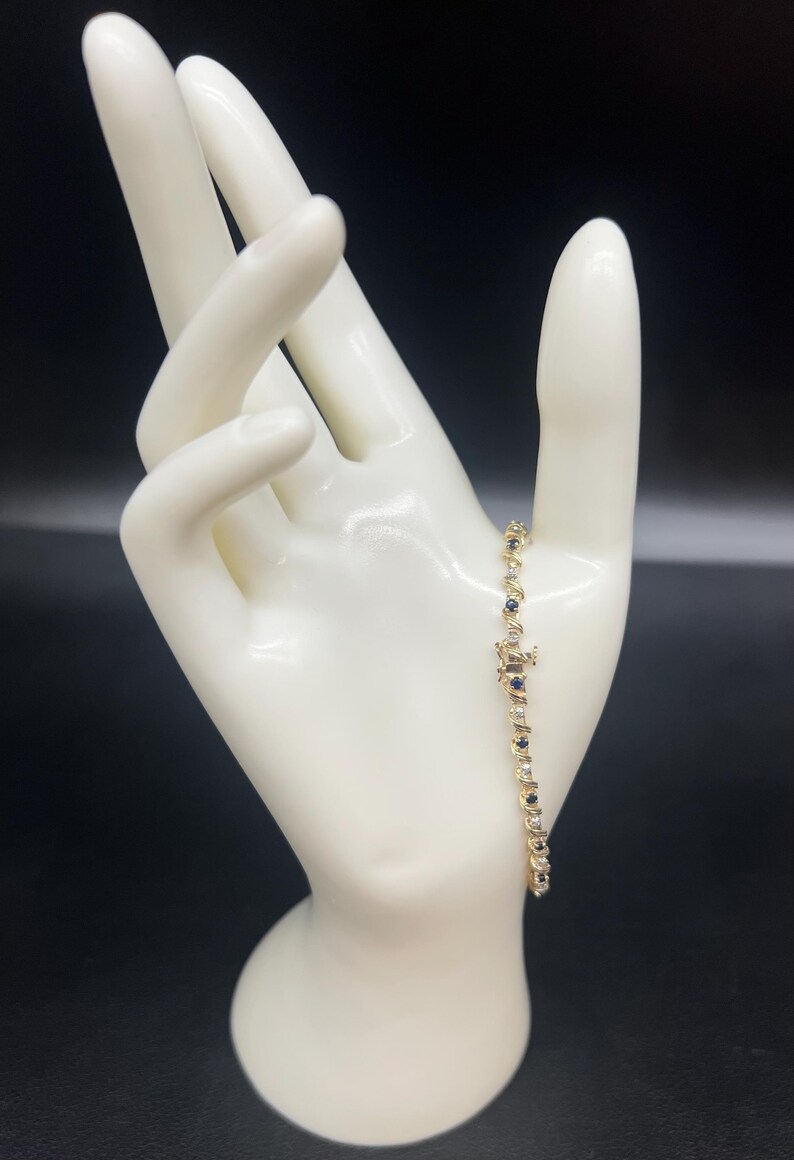 May include: A gold bracelet with alternating blue and white gemstones. The bracelet is on a white mannequin hand.