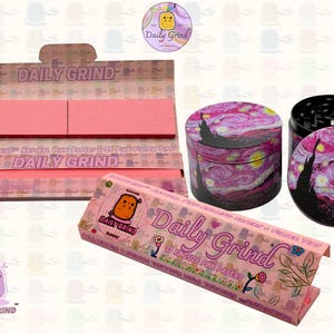 A pink and white box with the text 'Daily Grind' and ‘Kingsize Pink Papers' on it. The box is open and there are Pink papers inside. There is a tobacco grinder next to the box with a pink night sky art design and The 'Daily Grind' logo is in the corner of the image