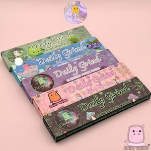 Puede incluir: Cinco paquetes rectangulares de papel de fumar Daily Grind apilados. Los paquetes presentan diseños en verde, azul, morado, rosa y negro con ilustraciones de dibujos animados, motivos florales y texto que indica 33 papeles king size.
