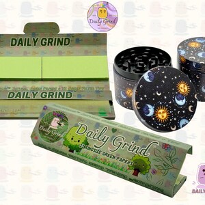 A dark Purple packet and a Green box with the text 'Daily Grind' on both and ‘Kingsize Purple Papers' on one and ‘Kingsize Green Papers’ on the other. Both boxes open showing green papers in one and purple in the other. There is a tobacco grinder with a cartoon sun and moon astrology design on it underneath it and The 'Daily Grind' logo is in the corner of the image