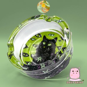 Puede incluir: Un cenicero de cristal redondo con un gráfico central de un gato negro rodeado de ojos verdes sobre un fondo verde lima. El exterior del cenicero muestra un patrón morado oscuro con pequeñas estrellas y el texto Daily Grind.