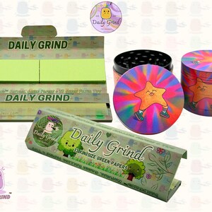 A dark Purple packet and a Green box with the text 'Daily Grind' on both and ‘Kingsize Purple Papers' on one and ‘Kingsize Green Papers’ on the other. Both boxes open showing green papers in one and purple in the other. There is a tobacco grinder with a very happy and confident star character in the middle with rays of light behind him underneath it and The 'Daily Grind' logo is in the corner of the image