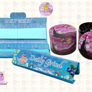 A deep blue and light blue box with the text 'Daily Grind' and ‘Kingsize Blue Papers' on it. The box is open and there are blue papers inside. There is a tobacco grinder with a cute pink art design on it next to the box and The 'Daily Grind' logo is in the corner of the image