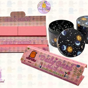 A pink and white box with the text 'Daily Grind' and ‘Kingsize Pink Papers' on it. The box is open and there are Pink papers inside. There is a tobacco grinder with a cartoon sun and moon astrology design on it next to the box and The 'Daily Grind' logo is in the corner of the image