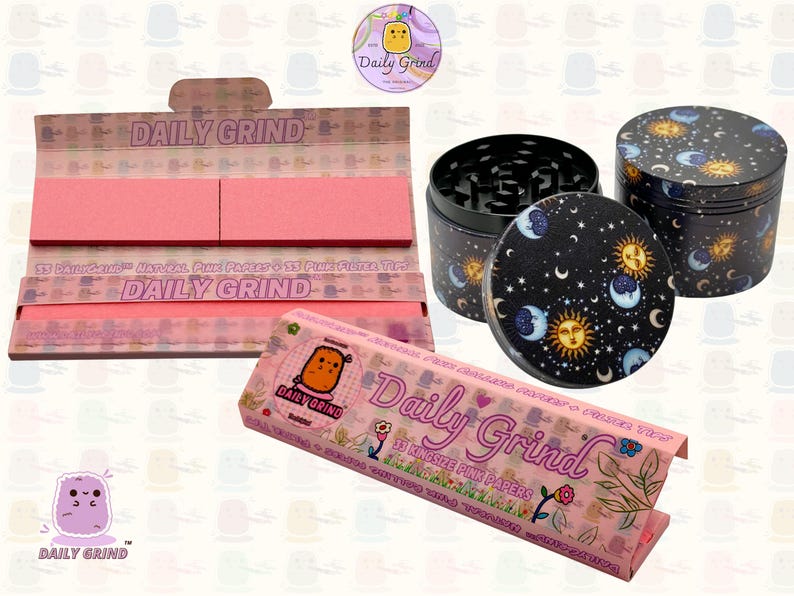 A pink and white box with the text 'Daily Grind' and ‘Kingsize Pink Papers' on it. The box is open and there are Pink papers inside. There is a tobacco grinder with a cartoon sun and moon astrology design on it next to the box and The 'Daily Grind' logo is in the corner of the image
