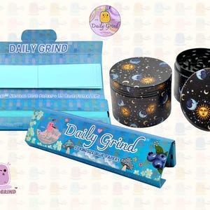 A deep blue and light blue box with the text 'Daily Grind' and ‘Kingsize Blue Papers' on it. The box is open and there are blue papers inside. There is a tobacco grinder with a cartoon sun and moon astrology design on it next to the box and The 'Daily Grind' logo is in the corner of the image