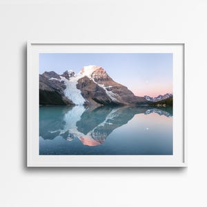 May include: A framed landscape photograph of a mountain reflected in a calm lake. The mountain has a snow-covered peak and a glacier. The sky transitions from pink to blue, and a moon is visible. The water is a light blue color.