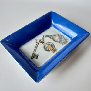 May include: A rectangular, blue-rimmed ceramic dish with a white interior. The dish features a gold key and a decorative tassel design. The key has a tag that reads "Imperial".