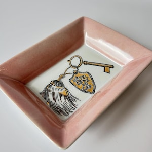 May include: A rectangular, pink ceramic dish with a white interior. The dish features a gold key and a tag that reads "Hôtel Côte du Cap" with a gold tassel. The dish is likely for holding small items.