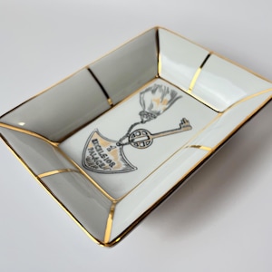 May include: Rectangular white and gray ceramic tray with gold trim. The tray features a central design of a key and a shield with the text "Excelsior Palace". A decorative item.