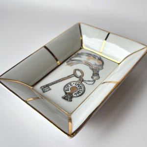 May include: A rectangular white ceramic trinket dish with gold trim and a decorative key and tassel design. The dish features a black and gold illustration of a key and a tassel with the words "Ritz Imperial" on the key's fob.