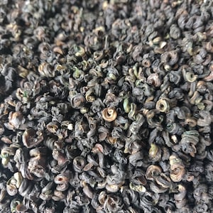 May include: Close-up of a pile of dark green tea leaves, tightly curled into spirals. The tea leaves have a slightly glossy appearance, with hints of lighter green and brown tones visible throughout the mass. The texture appears dense and compact.