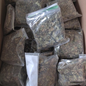 May include: A box filled with clear plastic bags containing dried green leaves. The bags are stacked on top of each other and some have labels that say "Products of Nigeria".