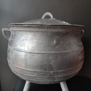 May include: A round, silver-colored metal pot with a textured, weathered finish. The pot features three short legs, two side handles, and a matching lid with a small central handle. The surface has a rough, cast appearance with horizontal bands.