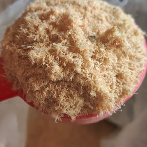 May include: Close-up of a red scoop filled with light brown, shredded material. The material appears to be finely shredded, with a textured, fibrous appearance. The scoop is held against a neutral background, highlighting the texture and color of the material.