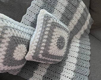 Könnte beinhalten: Eine graue und weiße gehäkelte Wolldecke mit einem gewellten Rand und zwei passenden gehäkelten Kissen. Die Decke und die Kissen liegen auf einem grauen Sofa.