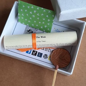 May include: A small white box with a green and white patterned paper inside. The box contains a rolled up scroll with an orange ribbon, a small coin, and a match. The scroll has the text "The Wish by Rhys Thomas" printed on it. The box is open and the scroll is partially unrolled.