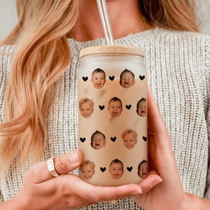 May include: A clear glass tumbler with a bamboo lid and a glass straw. The tumbler is decorated with smiling baby faces and black heart shapes. The tumbler is held by a person wearing a gray sweater.