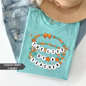 chalky mint Comfort Colors® shirt with colorful beaded bracelet design spelling "The Life Of A Teacher", surrounded by school-themed icons