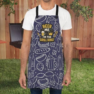 May include: Navy blue apron featuring a white food and beer design. The apron has the text "BEER ME, I'M THE GRILL KING!" in yellow and white. Black straps and buttons complete the design. Ideal for barbecues.