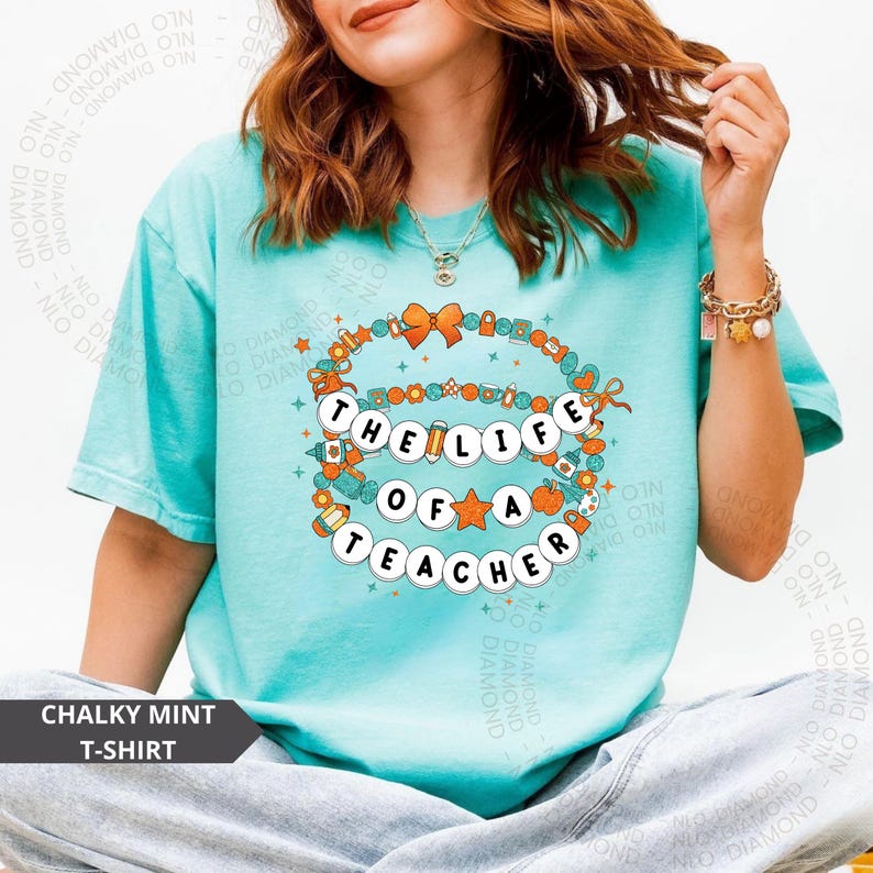 CHALKY MINT comfort colors shirt with orange handwritten text “the life of a teacher” and mini sparkles, worn by woman showing peace sign