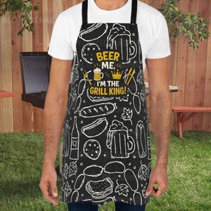 May include: Black apron with white food illustrations and the text "BEER ME, I'M THE GRILL KING!" in yellow. The apron features drawings of beer mugs, hot dogs, and burgers. The apron has black straps and is worn by a person.