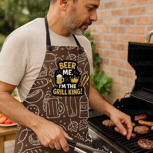 May include: Brown apron with a white food and beer-themed pattern. The apron features the text "BEER ME, I'M THE GRILL KING!" in yellow and black. A person is grilling burgers and sausages on a black barbecue.