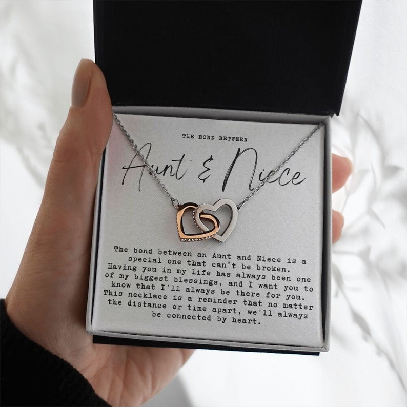May include: A silver necklace with two interlocking hearts, one rose gold and one silver. The necklace comes in a box with a message that reads "The bond between an Aunt and Niece is a special one that can't be broken. Having you in my life has always been one of my biggest blessings, and I want you to know that I'll always be there for you. This necklace is a reminder that no matter the distance or time apart, we'll always be connected by heart."