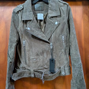 May include: A brown suede jacket with a classic biker style. Features include an asymmetrical zipper closure, multiple zippered pockets, a belted waist, and shoulder epaulets. The jacket is hanging on a black hanger against a wooden door.