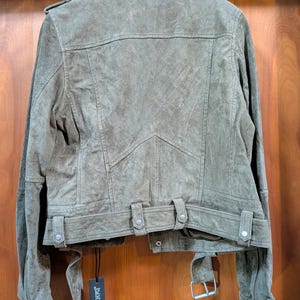 May include: Olive green suede jacket with a classic biker style. Features include a notched collar, shoulder epaulets, zippered sleeves, and a belted waist. The jacket has a textured finish and a tag that reads "BLANKNYC".