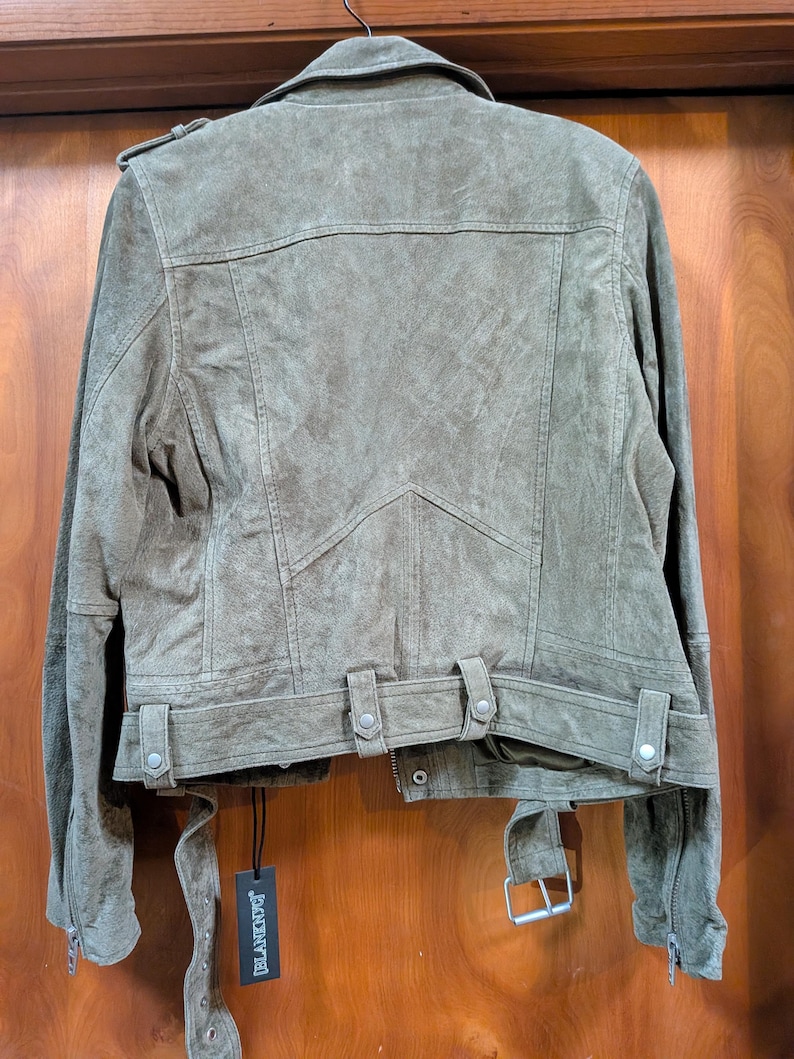 May include: Olive green suede jacket with a classic biker style. Features include a notched collar, shoulder epaulets, zippered sleeves, and a belted waist. The jacket has a textured finish and a tag that reads "BLANKNYC".