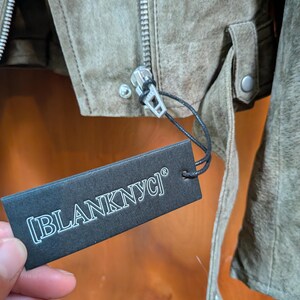 May include: Close-up of a suede jacket in a muted olive green color. The jacket features a silver zipper, a small strap with a button, and a tag that reads "[BLANKNYC]" in white text.